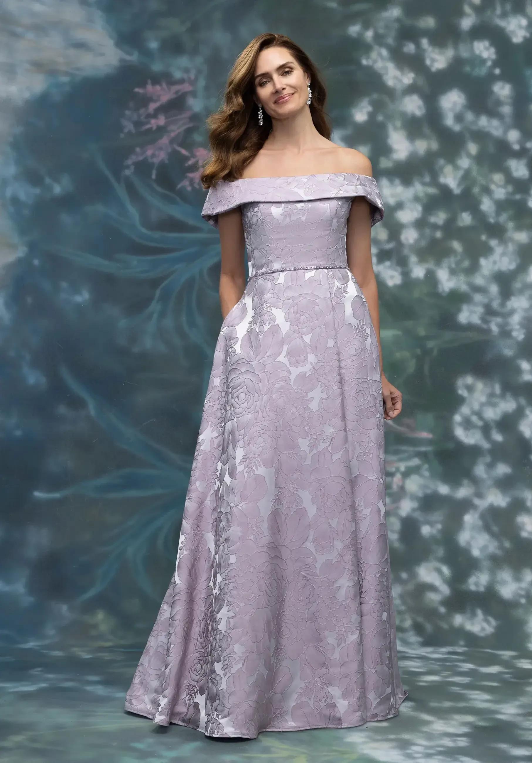 A woman wearing an elegant lavender off-the-shoulder gown with floral patterns, standing against a soft, blurred background.