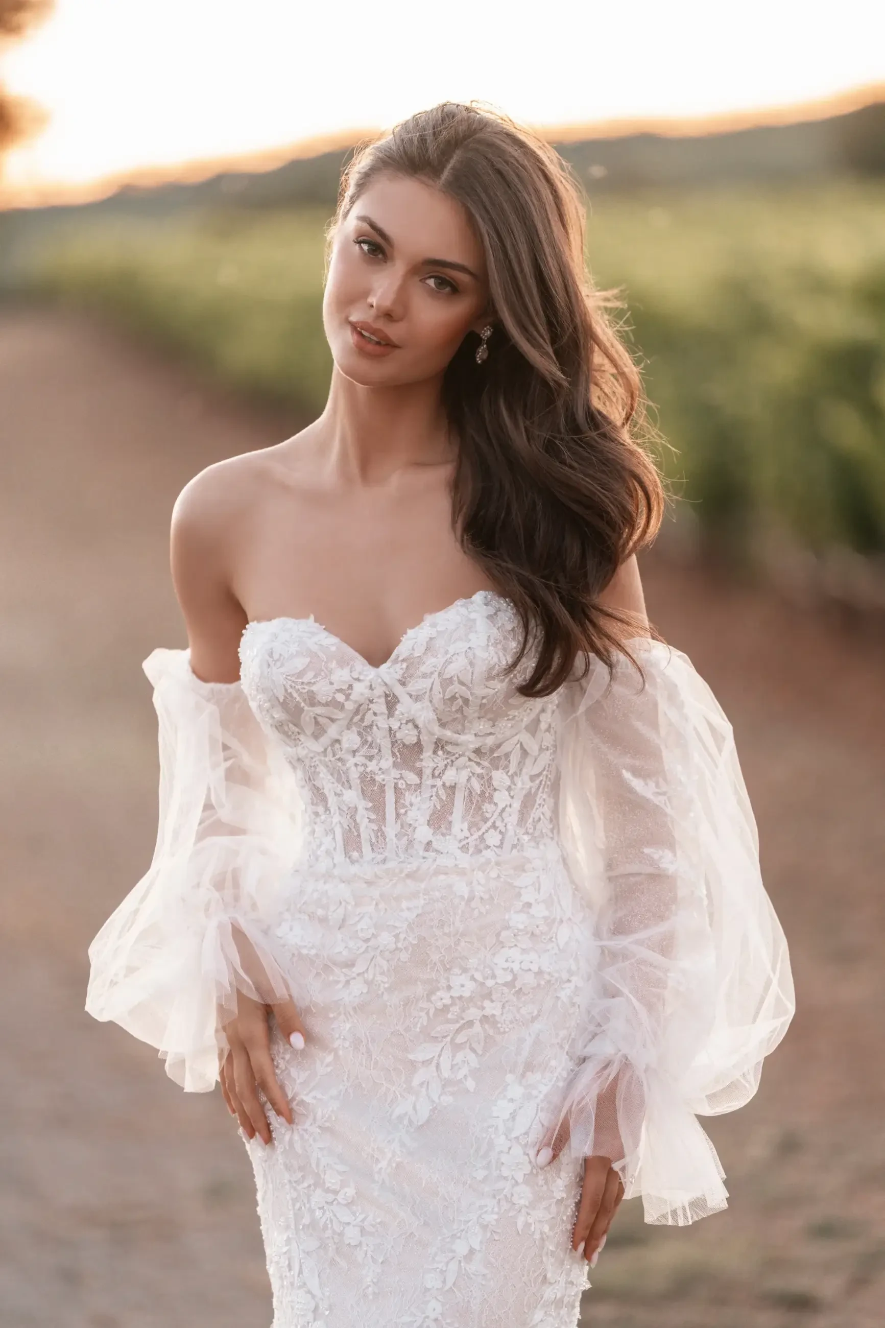 A woman in a lace wedding dress with sheer sleeves stands on a sunlit path. Her long hair cascades, conveying a serene and romantic tone.