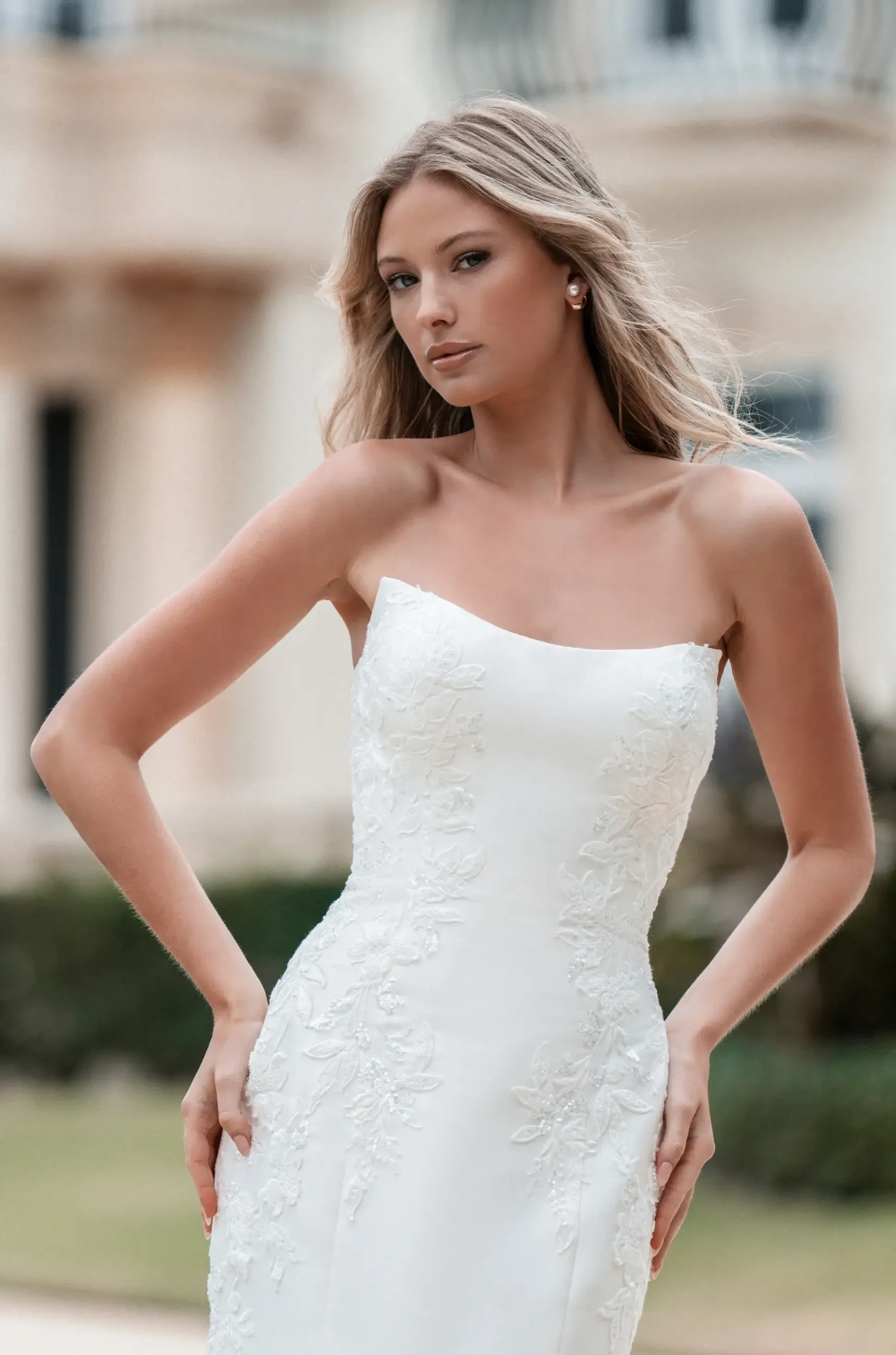 A woman in a strapless white wedding dress with lace details poses confidently outdoors. Her long hair is styled naturally, conveying elegance and grace.