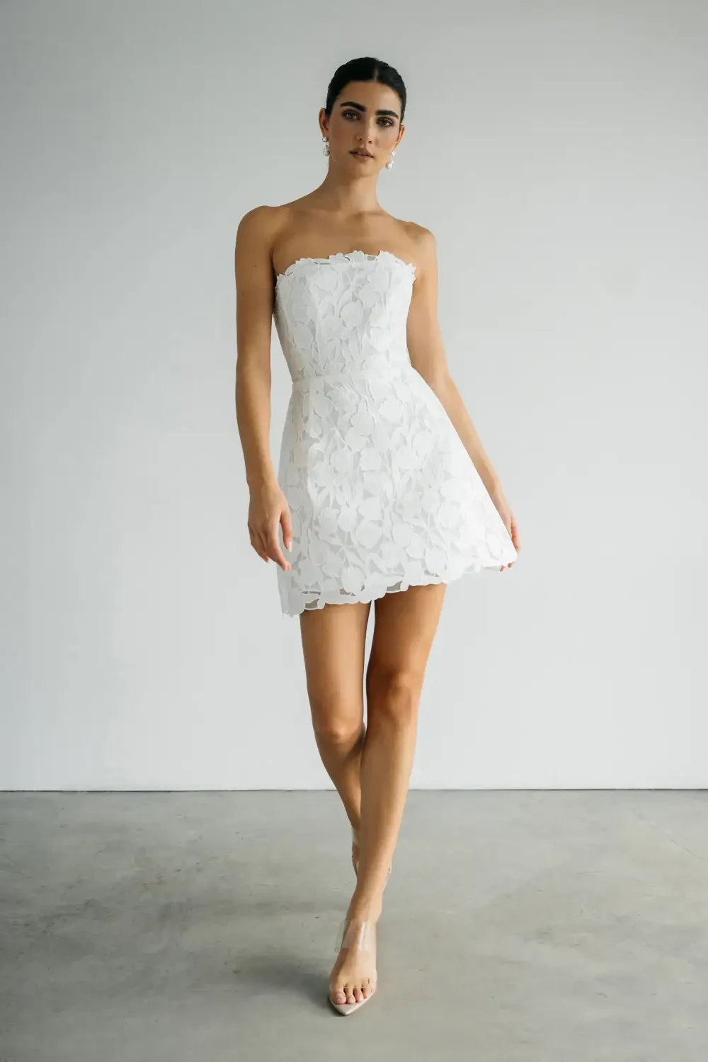A model wearing a strapless white dress standing in a minimalistic setting.