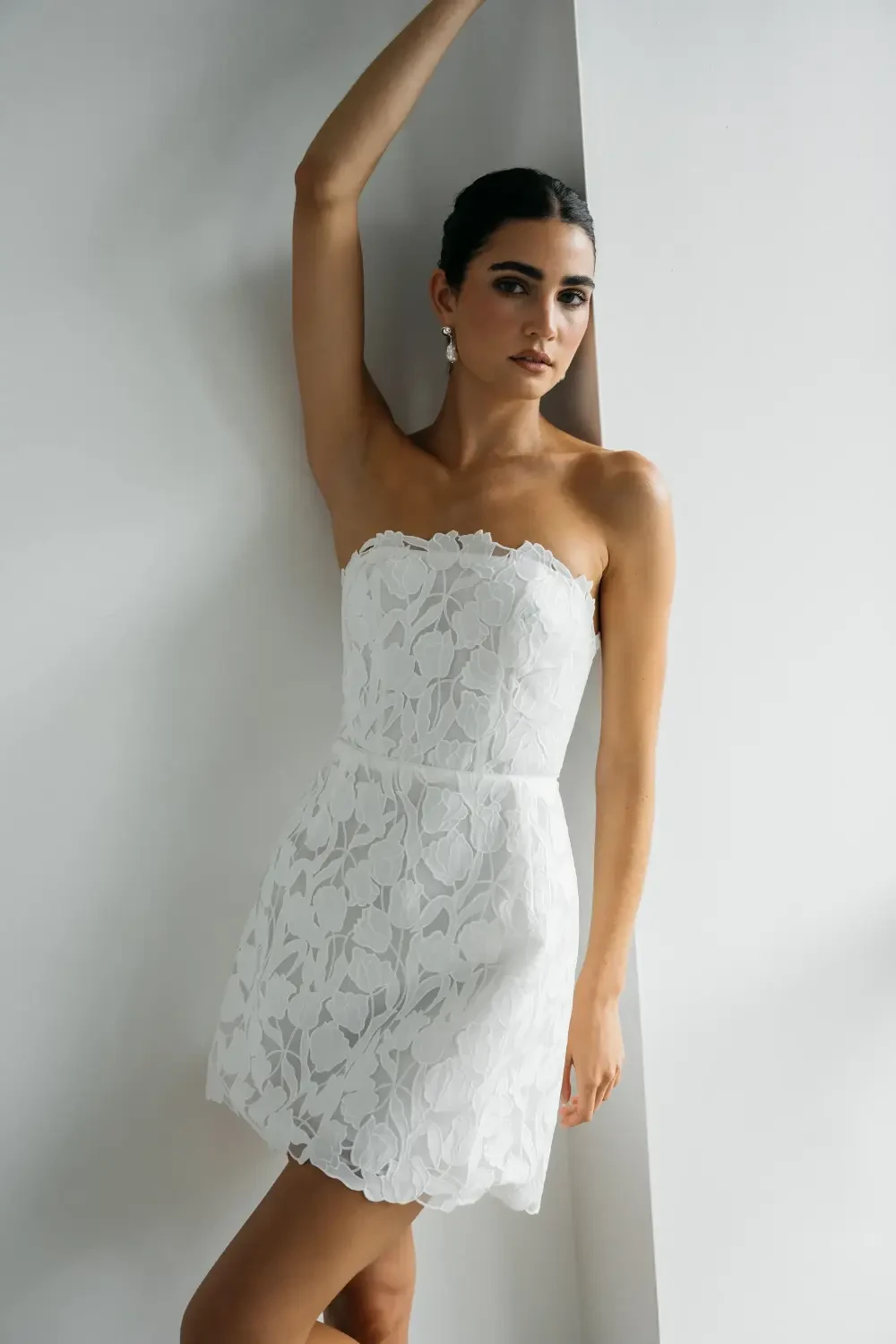 A model wearing a strapless white dress standing in a minimalistic setting.