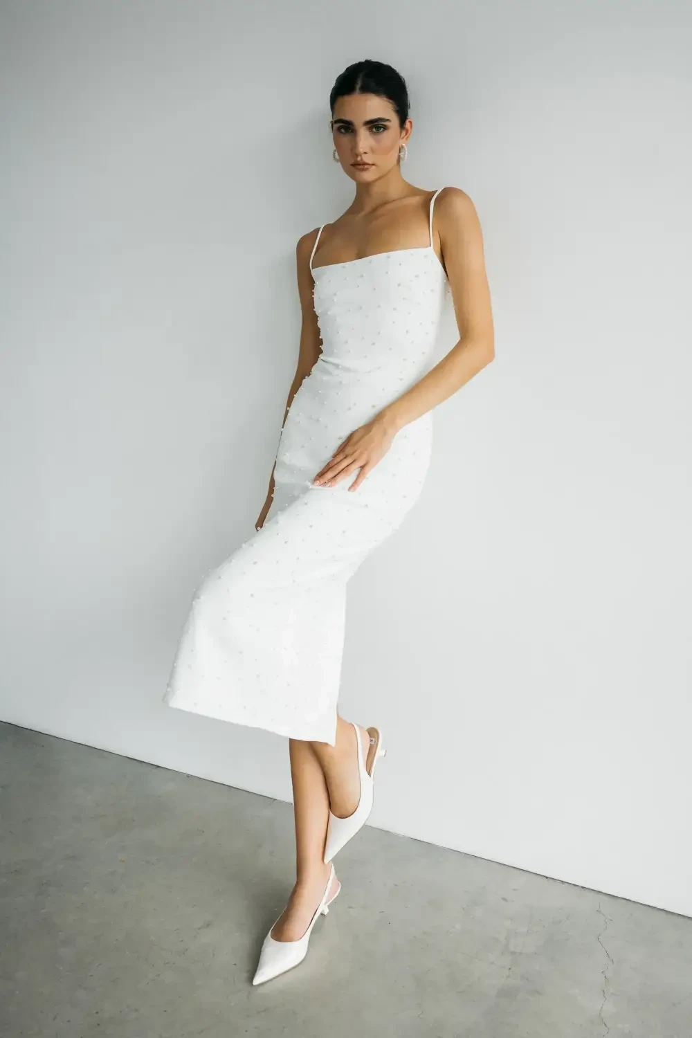 A woman poses in a fitted white dress with spaghetti straps and pointed-toe heels against a minimalistic background.