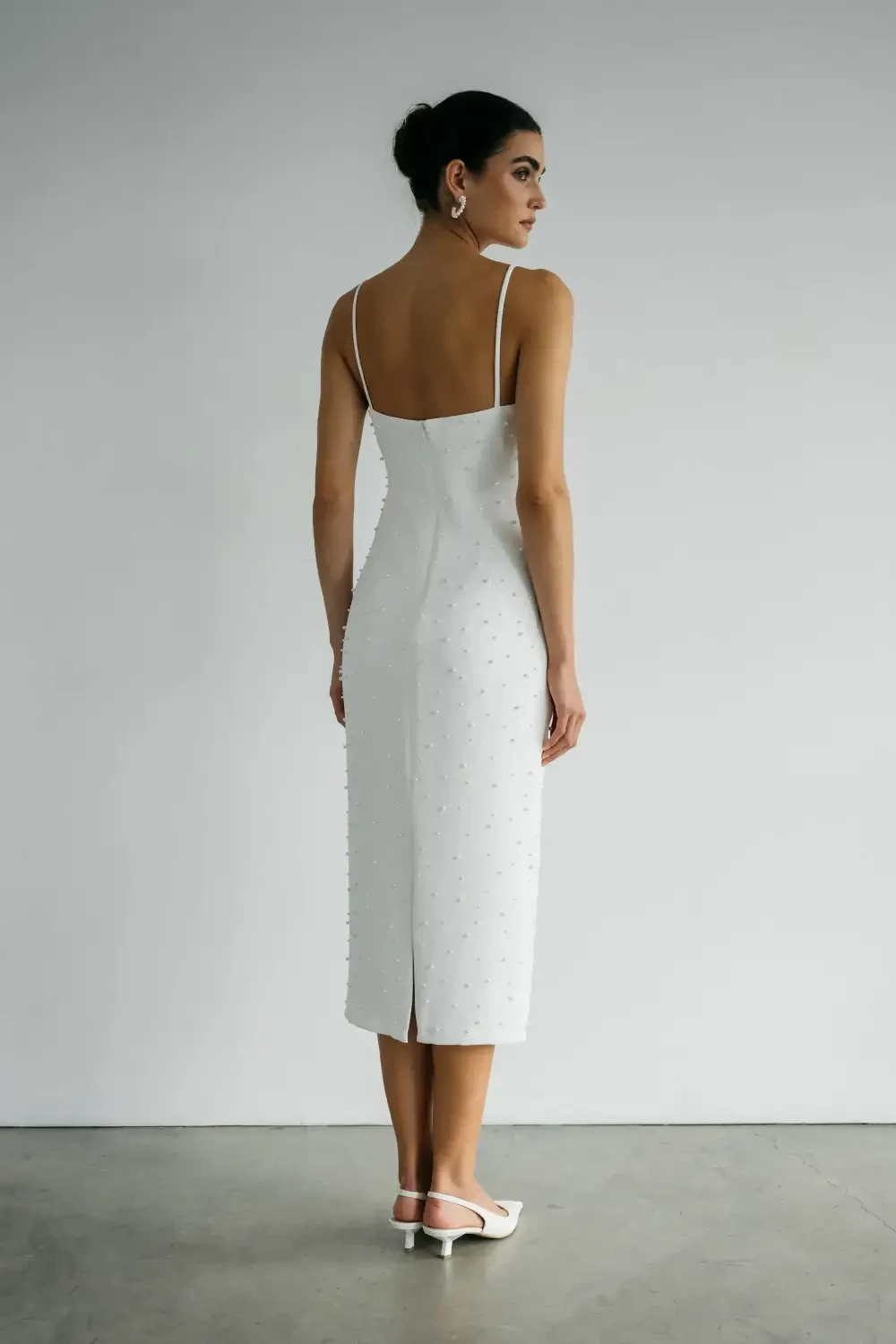A woman poses in a fitted white dress with spaghetti straps and pointed-toe heels against a minimalistic background.