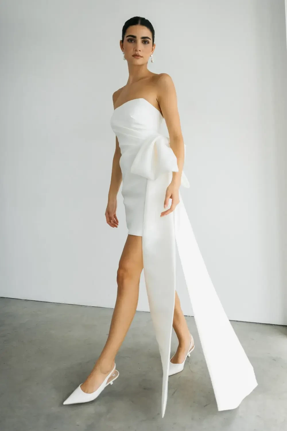 A model walking in a short white strapless dress with a large bow and long trailing fabric, paired with white heels.