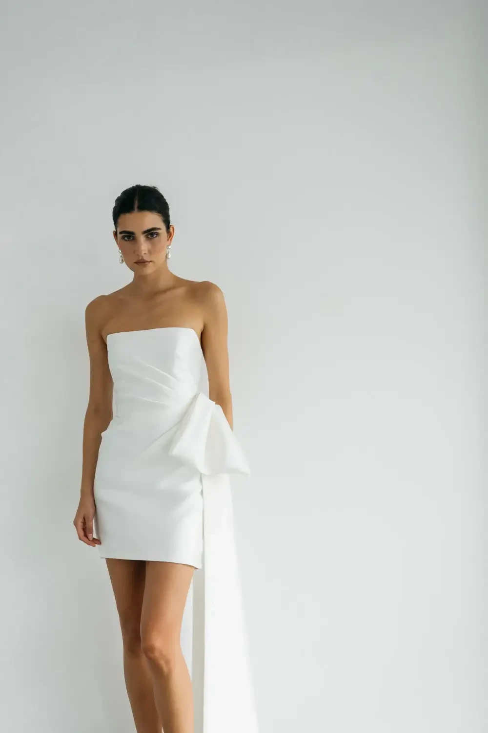 A model walking in a short white strapless dress with a large bow and long trailing fabric, paired with white heels.